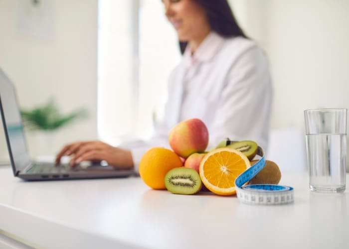 Nutricionista em consultório preparando dicas no notebook em mesa com frutas, copo de água e fita métrica